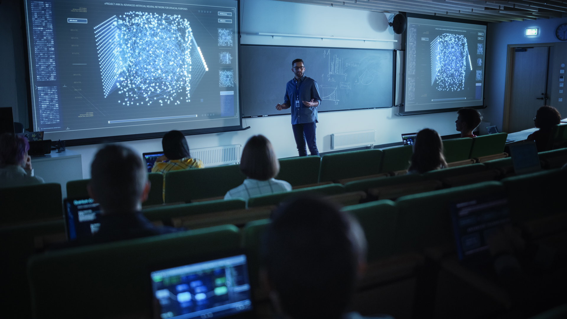 A color photo of a professor instructing students in a dark lecture hall